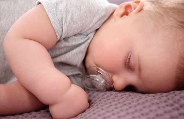 Baby mit einem durchsichtigen Schnuller im Mund schläft seitlich auf einer lila Decke.