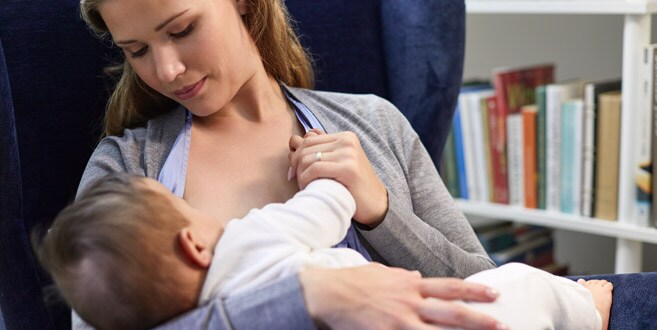 Eine Mutter lehnt sich in einem blauen Samtstuhl zurück und schaut auf ihr stillendes Baby hinunter, was den Wiegengriff demonstriert.