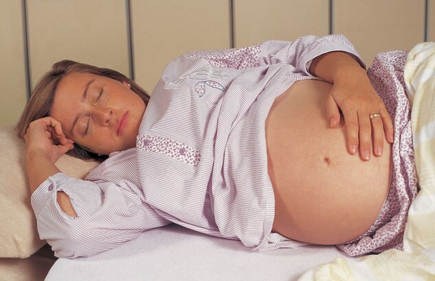 Hochschwangere Frau schläft auf der Seite mit einer Hand auf dem Bauch und der anderen beim Gesicht.