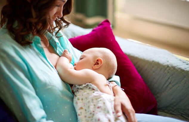 Mutter in einer blauen Bluse stillt ihr Baby sitzend auf einem bequemen grauen Sofa.