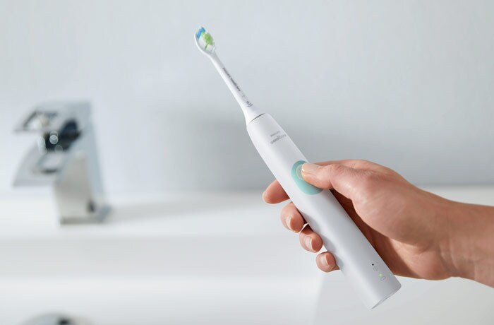Eine Hand, die eine weiße elektrische Philips Sonicare-Zahnbürste über ein Waschbecken hält und mit dem Daumen auf dem Knopf drückt.