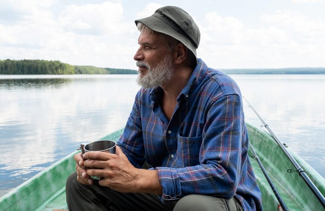 Ein älterer Mann mit grauem Bart, Fischerhut und blau kariertem Hemd sitzt auf einem Boot und blickt in die Ferne.