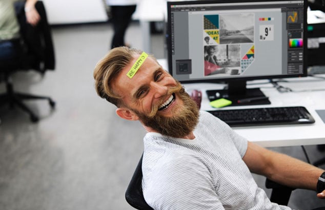 Ein Mann mit einem Hipster-Bart und einem Post-it mit dem Schriftzug „Be happy“ auf der Stirn, sitzt in einem Büro.