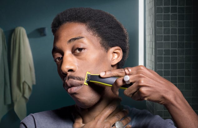 Ein junger, schwarzer Mann trimmt mit dem Philips OneBlade seinen Schnurrbart vor einem Spiegel in einem Badezimmer.